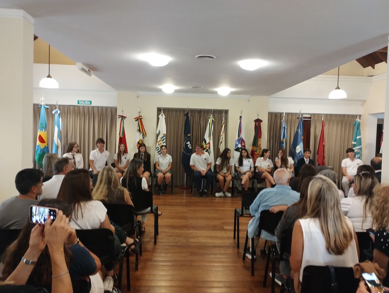 Ceremonia de la Medalla al Mérito: estudiantes sentados al frente, rodeados de banderas, y las familias en el público